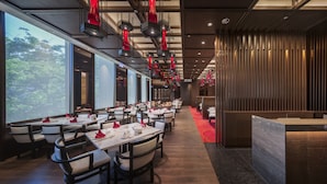 Un restaurante moderno con mesas largas, luces colgantes rojas y vistas a árboles a través de grandes ventanales.