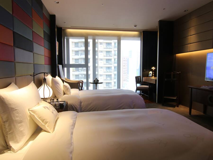 A hotel room with two beds, a TV, a desk, and a window with a city view.