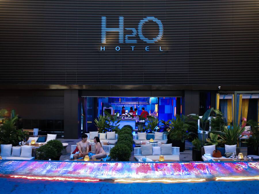A hotel entrance with a sign reading 'H2O HOTEL,' outdoor seating with cushions and tables, and potted plants.