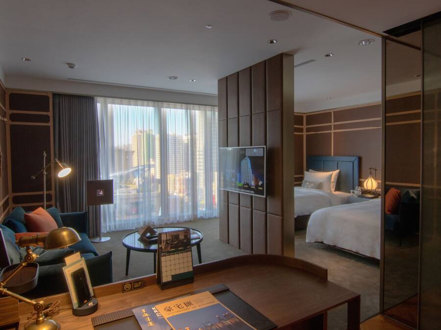 A hotel room with a bed, a desk with a lamp, a TV, and a view of the city through large windows.