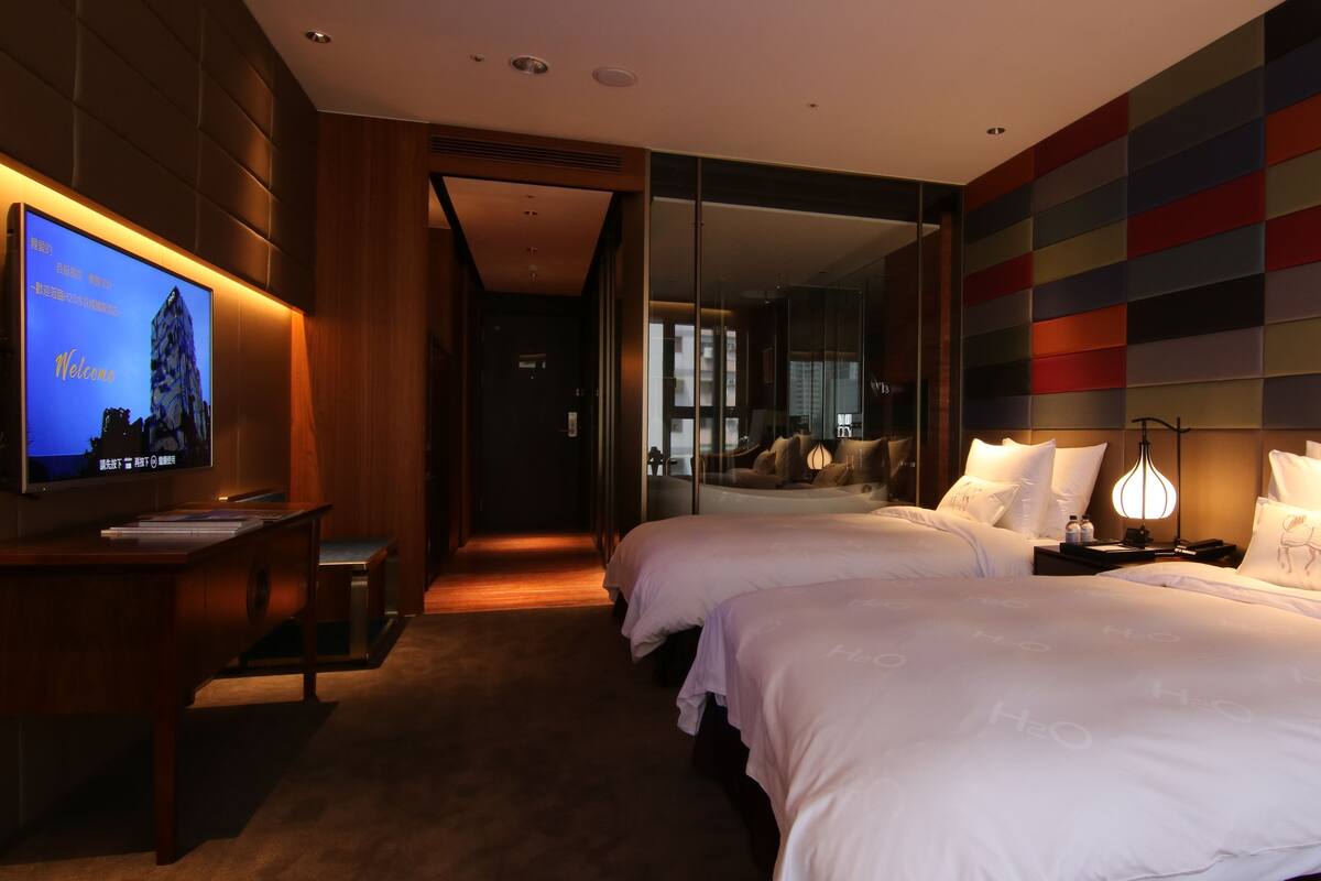 A hotel room with a large TV on the left, a bed with white linens, a bedside table with a lamp, and a colorful wall panel.