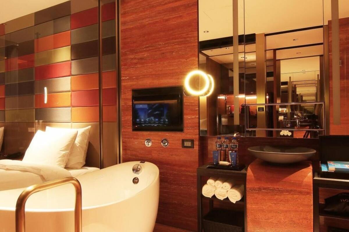 A modern hotel room with a large bed, a TV mounted on the wall, a round mirror, and a small table with toiletries.