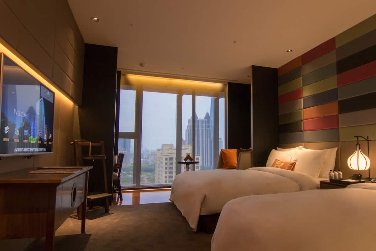 A hotel room with two beds, a TV, a desk, and a large window with a city view.