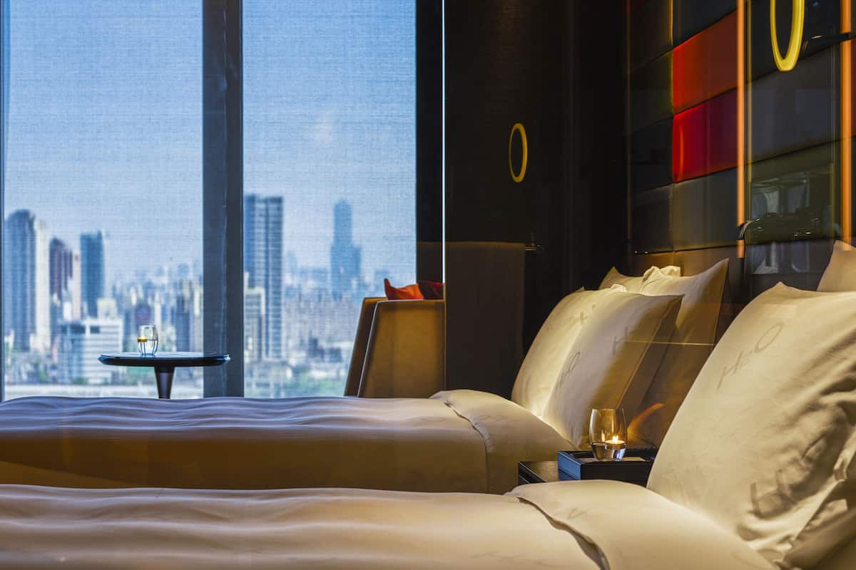 A hotel room with a large bed, a nightstand, and a view of a city skyline through large windows.