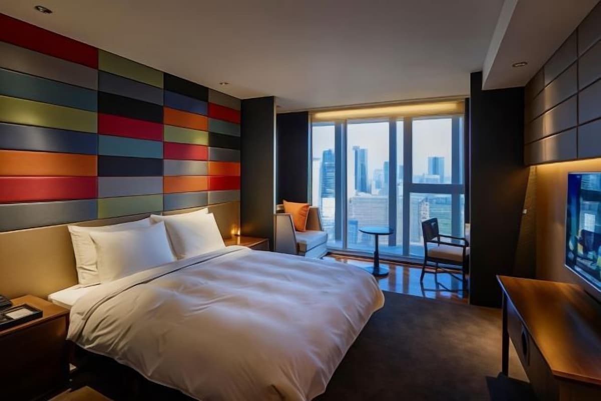A hotel room with a large bed, a desk, a chair, a TV, and a view of the cityscape through large windows.
