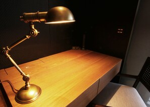Una lámpara de escritorio con acabado metálico sobre un escritorio de madera en una habitación con pared oscura y un enchufe visible.