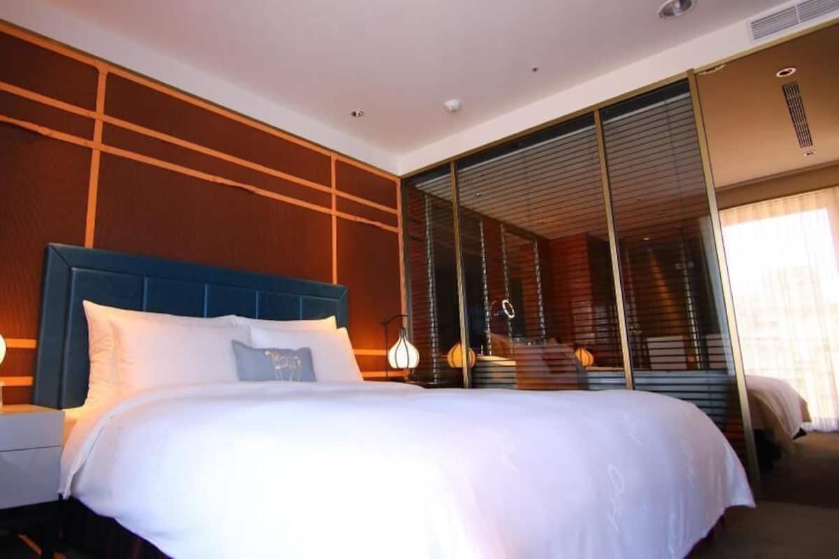 A hotel room with a large bed, a nightstand, a lamp, and a sliding glass door leading to a balcony.