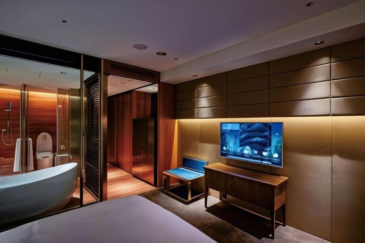 A modern hotel room with a large bed, a television, a bathroom with a bathtub, and a seating area.