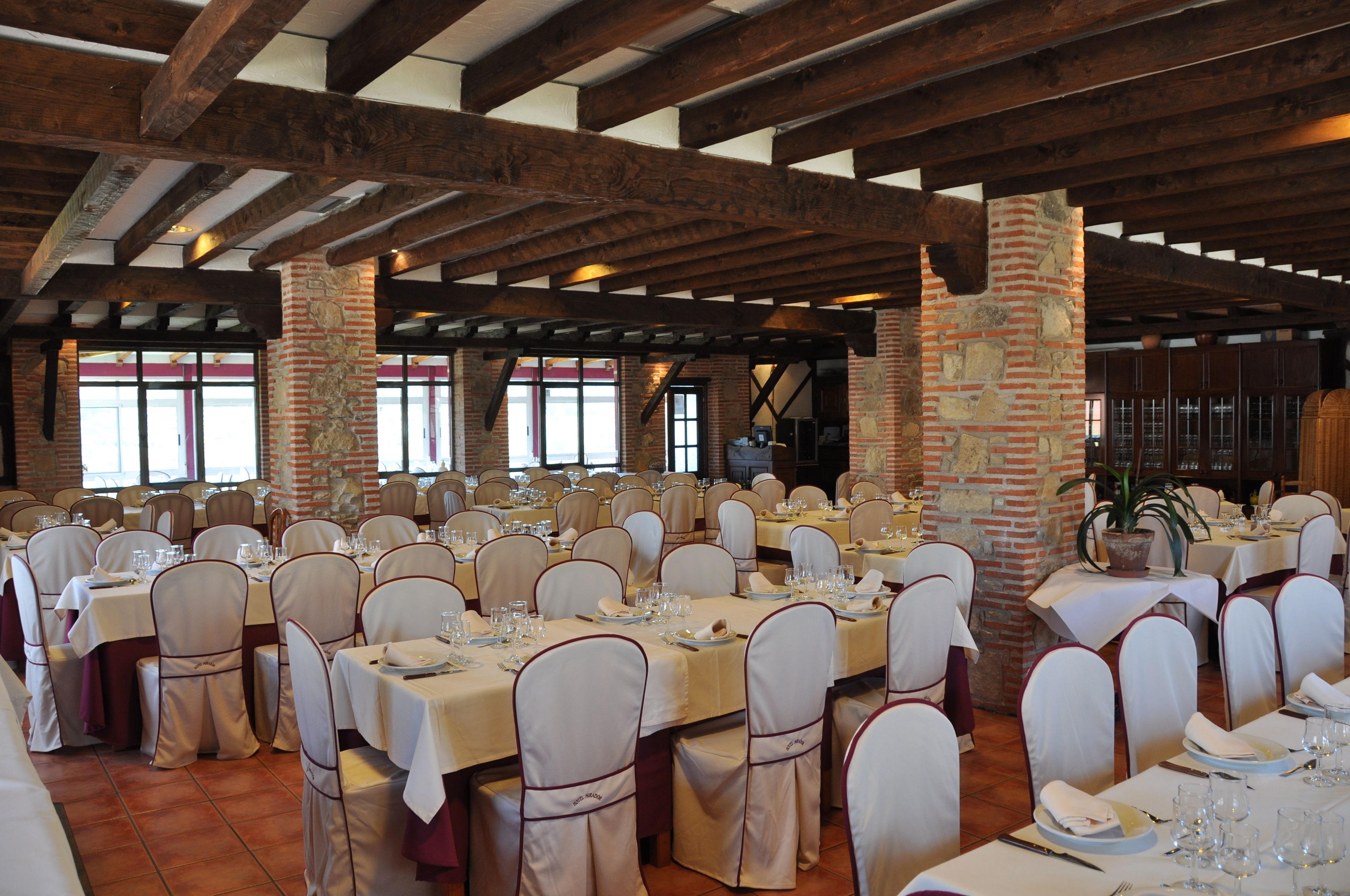 Un salón de banquetes con varias mesas dispuestas para comer, que cuenta con vigas de madera a la vista y pilares de ladrillo.