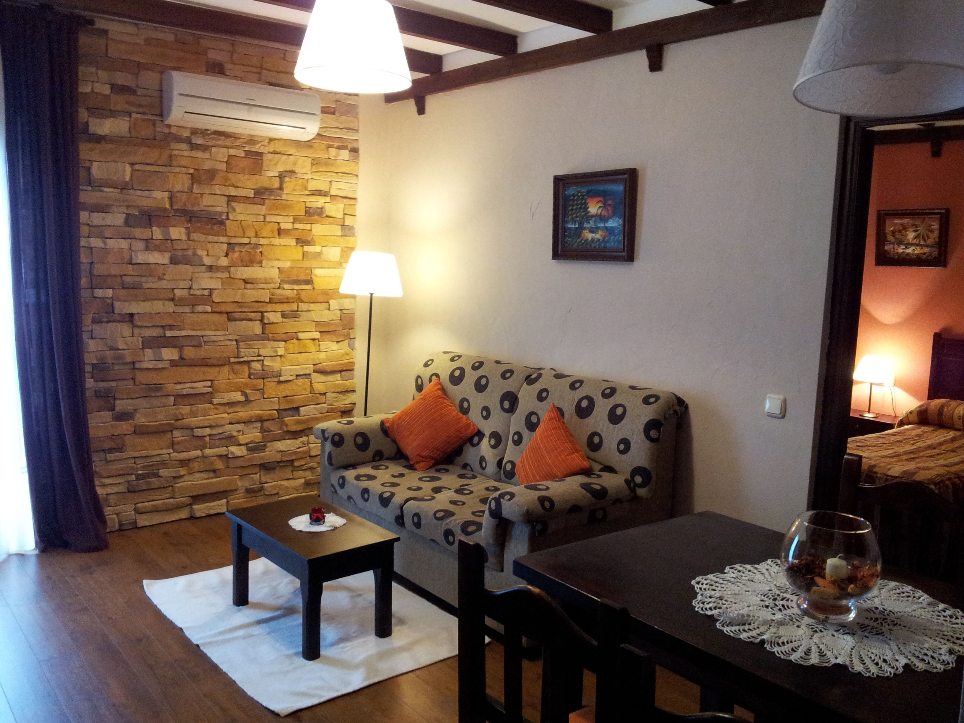 Een gezellige kamer met een stenen muur, een gestreept sofa met oranje kussens, een klein tafeltje met een glas en een witte kantklos.