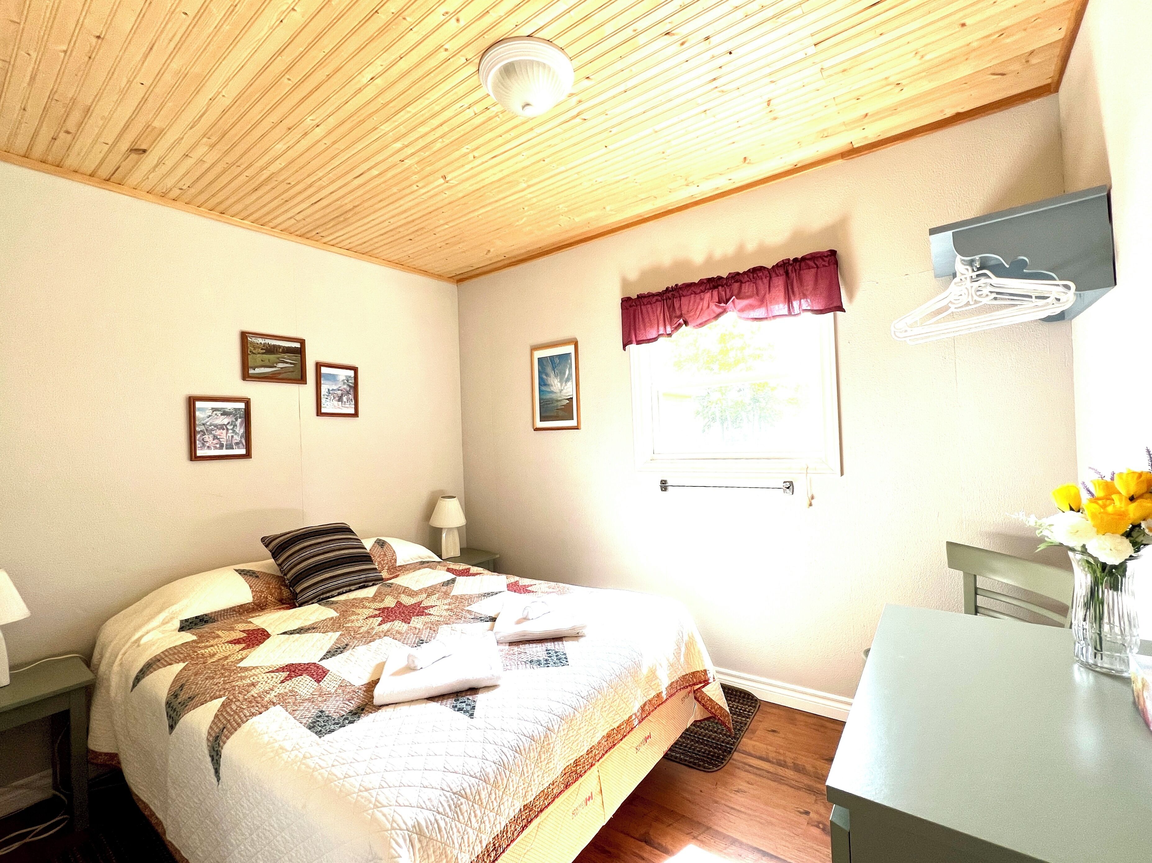 Un dormitorio con techo de madera, una cama con colcha, una ventana con cortina y un escritorio con un florero.