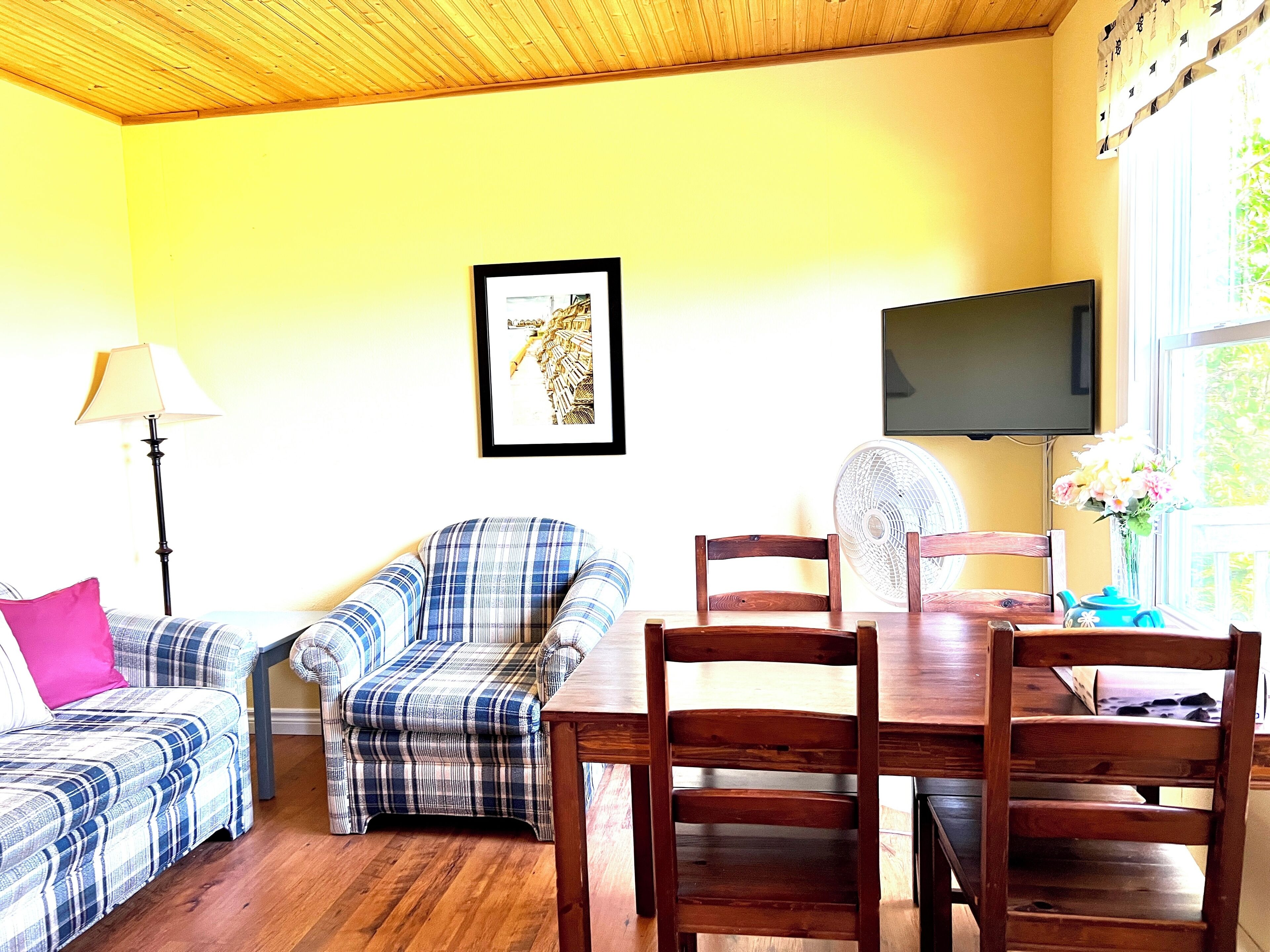 A living room with a wooden ceiling, a sofa, a chair, a dining table, a TV, a fan, and a window with flowers.