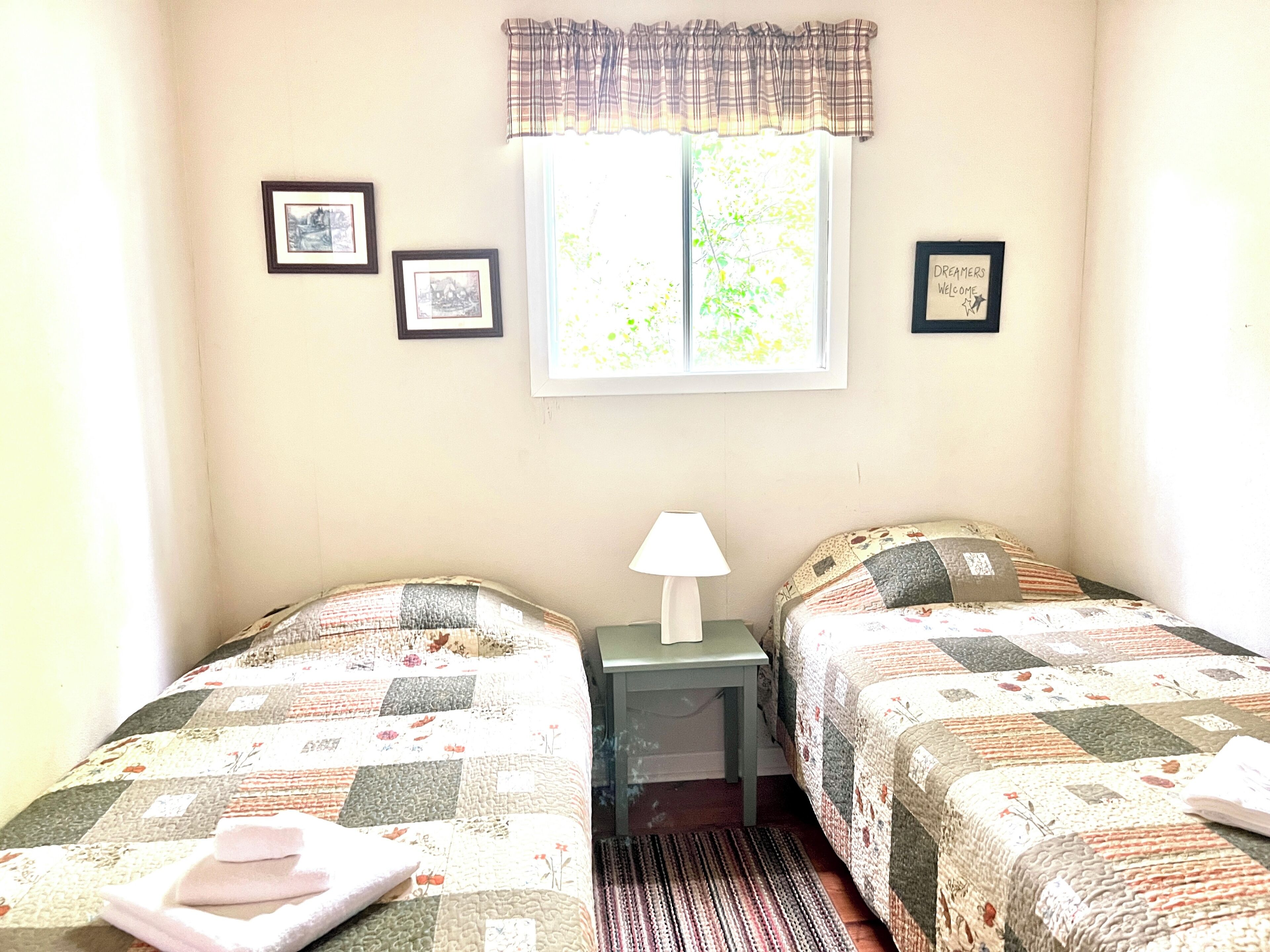 Ein Zimmer mit zwei Betten, einem fensterbelaubten Fenster, einem kleinen Tisch mit Lampe und gerahmten Bildern an der Wand.