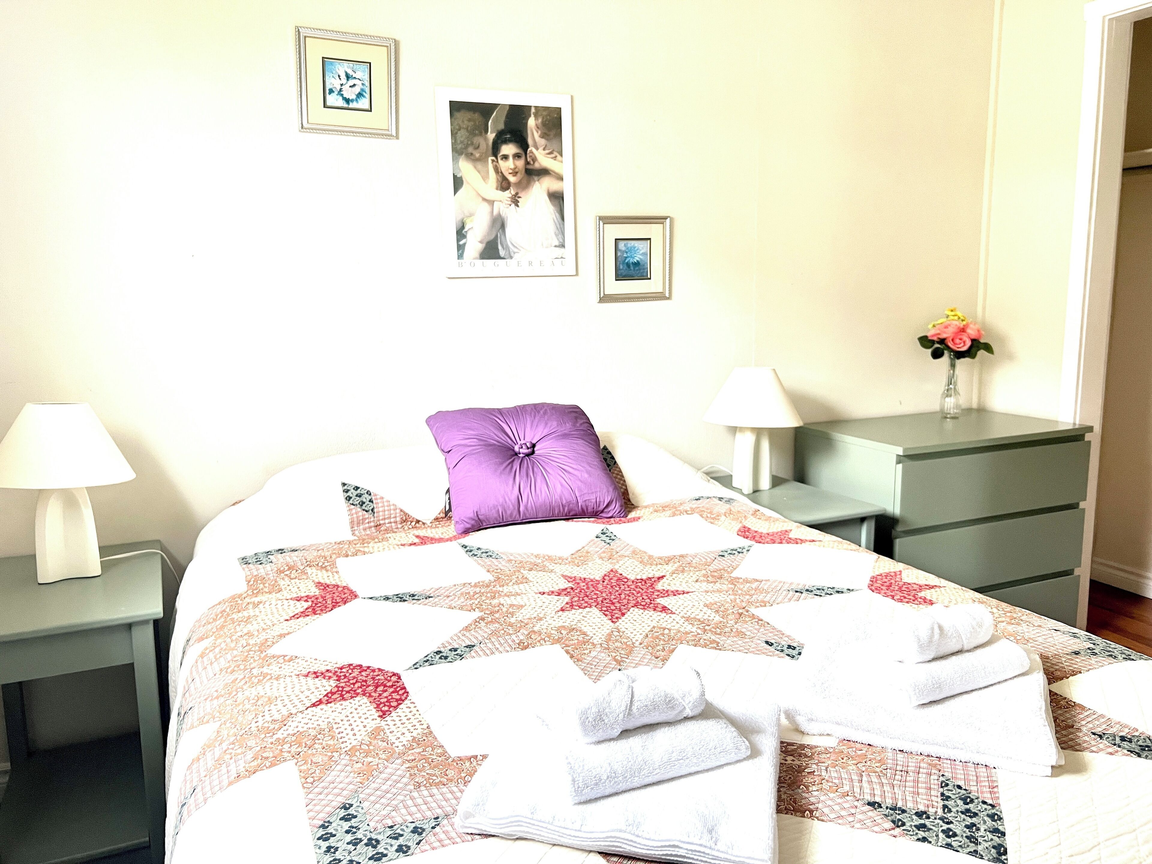A neatly made bed with a quilt, two bedside lamps, a purple pillow, and a small table with a vase of flowers.