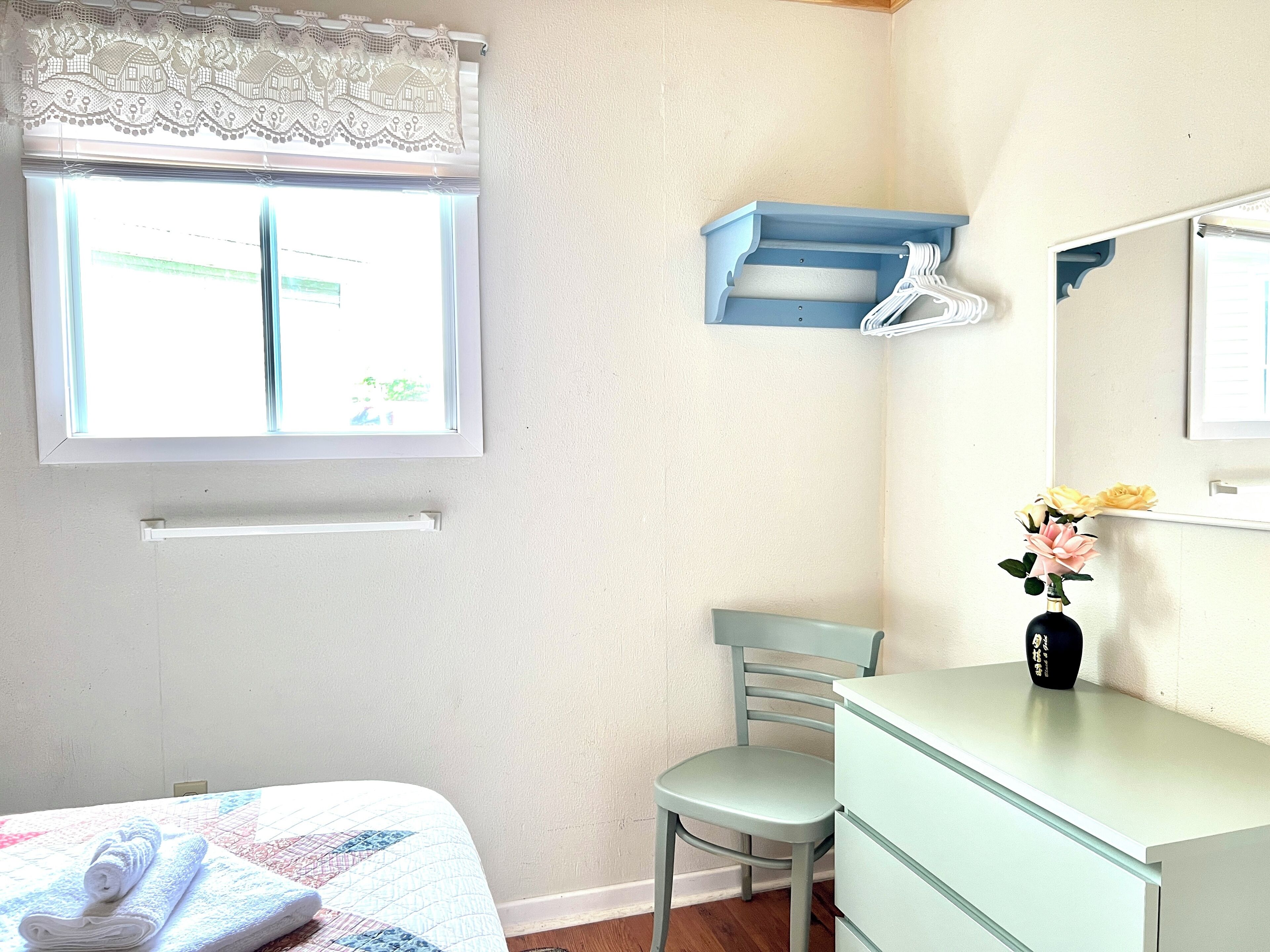 A bedroom with a bed, a chair, a dresser, a vanity with a mirror, and a window with a lace curtain.