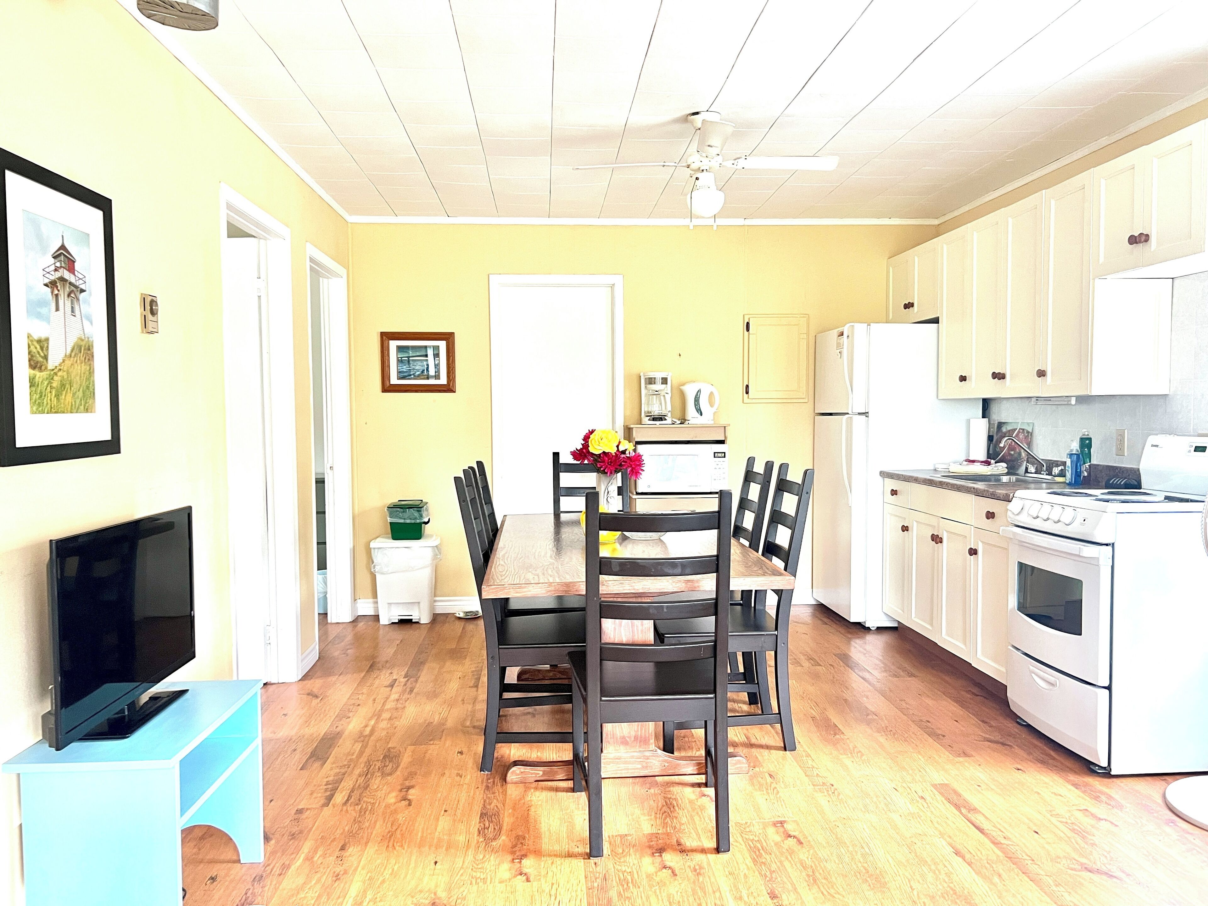 A bright kitchen with wooden cabinets, a dining table with chairs, a TV on a stand, and a ceiling fan.