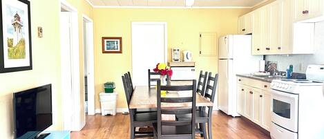 A bright kitchen with wooden cabinets, a dining table with chairs, a TV on a stand, and a ceiling fan.
