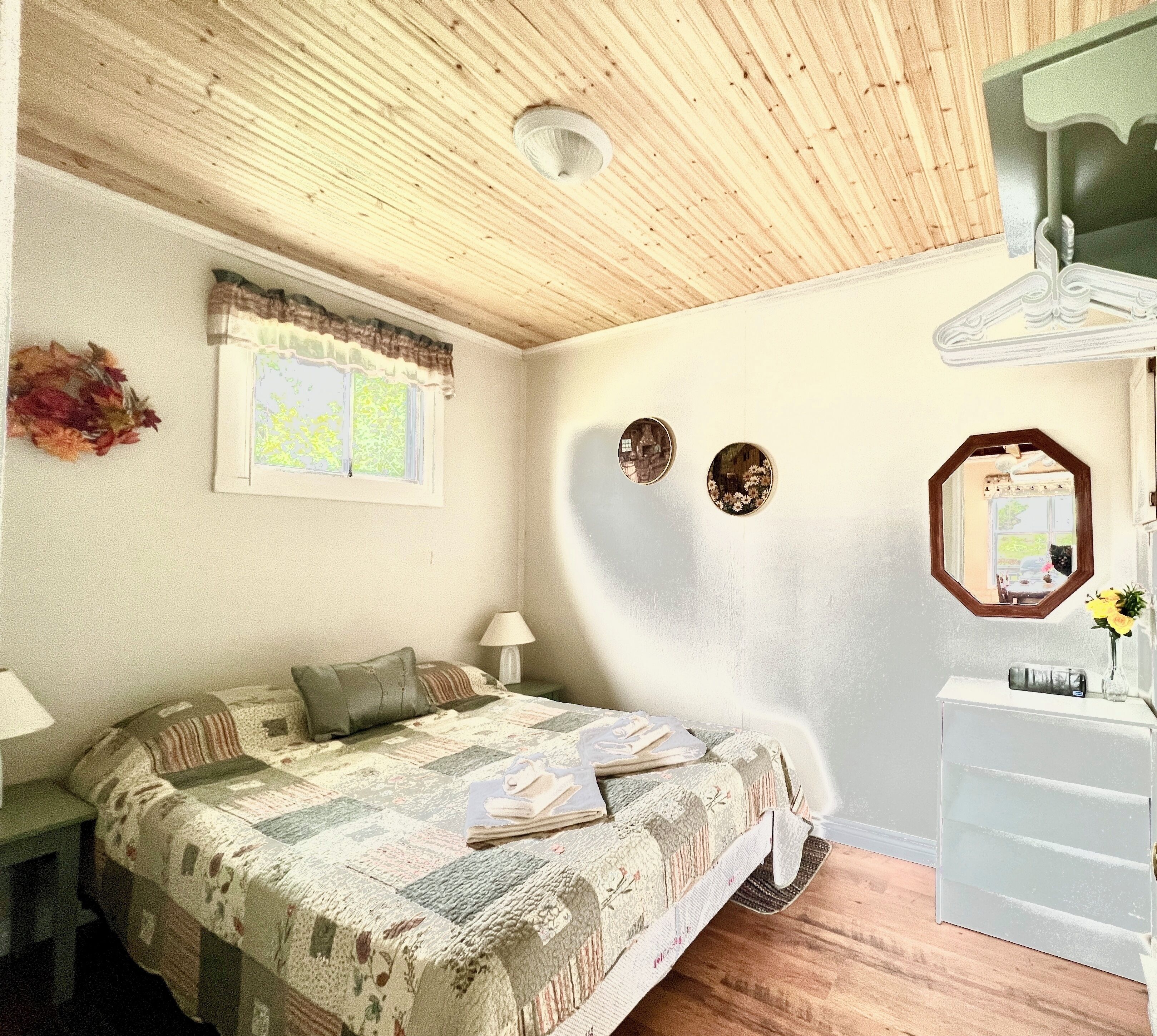 A bedroom with a wooden ceiling, a bed with a quilt, a nightstand, a lamp, a dresser, and a mirror.
