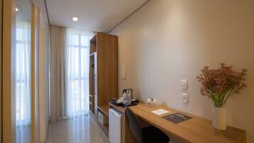 A modern hotel room with a wooden desk, a black office chair, a small refrigerator, and a vase with flowers on the desk.