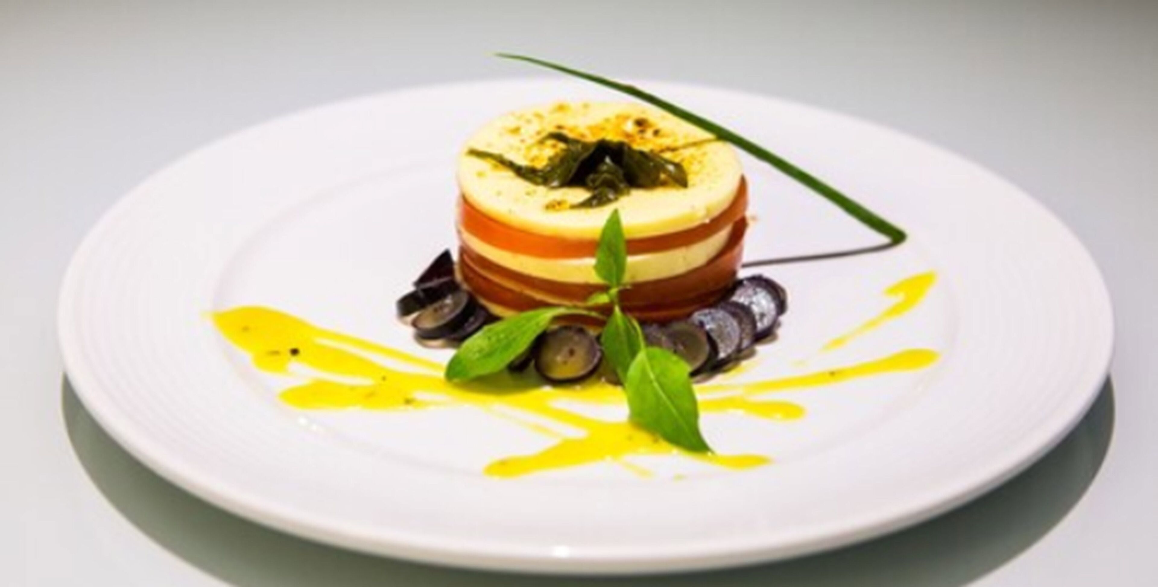 Um prato branco com um prato em camadas, possivelmente um terrine, decorado com uma folha verde e regado com molho amarelo.