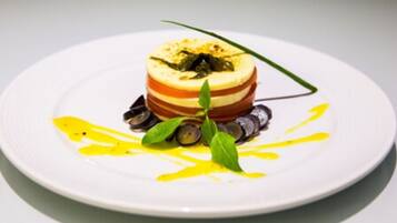 Um prato branco com um prato em camadas, possivelmente um terrine, decorado com uma folha verde e regado com molho amarelo.