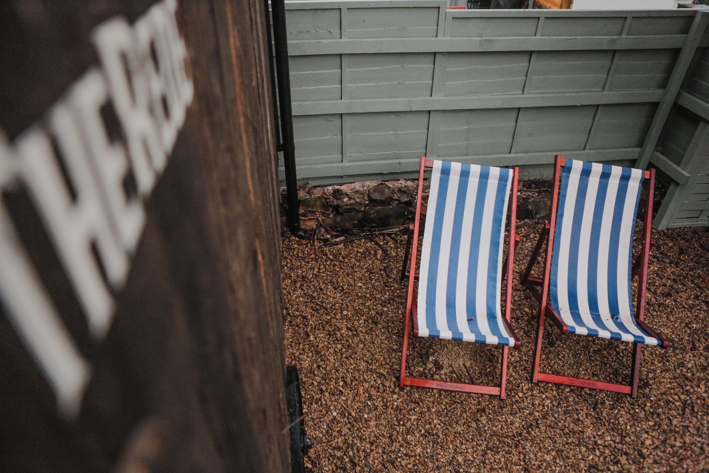Two striped beach chairs in a garden area with a wooden fence and a stone wall in the background.
