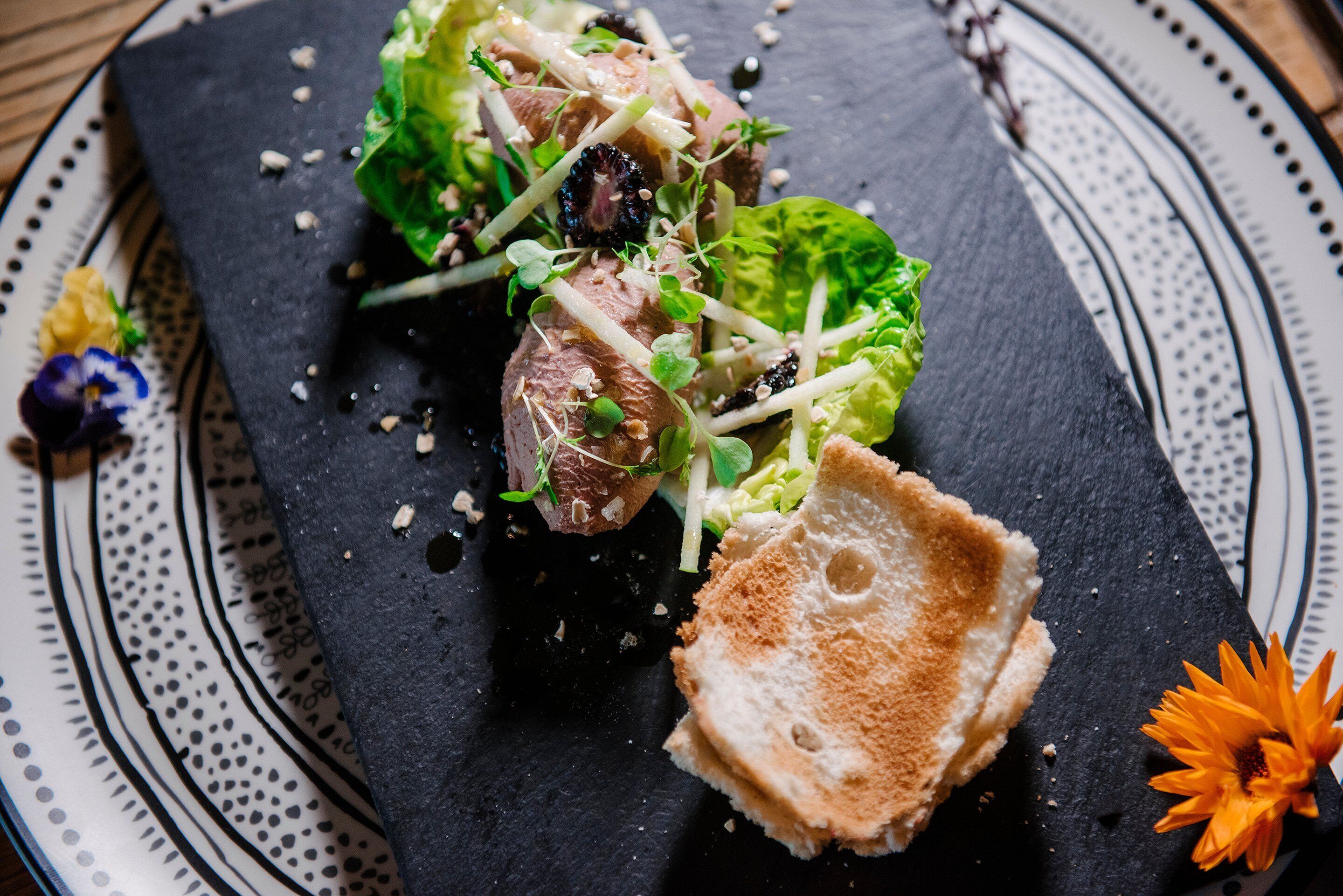 A black slate serving board with a salad, bread, and decorative flowers.