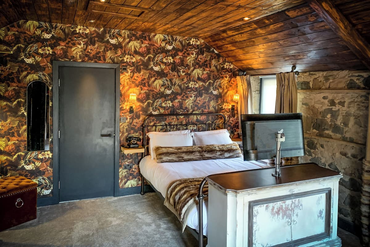 Une chambre avec un papier peint à motifs floraux, un lit recouvert de draps blancs, une table de chevet de style vintage et un mur en pierre.