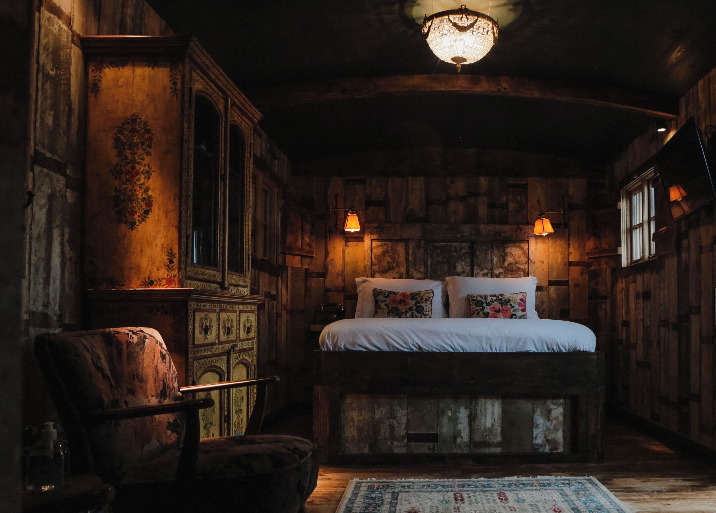 A rustic bedroom with a wooden headboard, a large bed with floral pillows, a wooden dresser, and a chandelier.