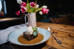 Un postre en un plato, con una cuchara a un lado, un jarrón floral al fondo y una superficie de mesa de madera.