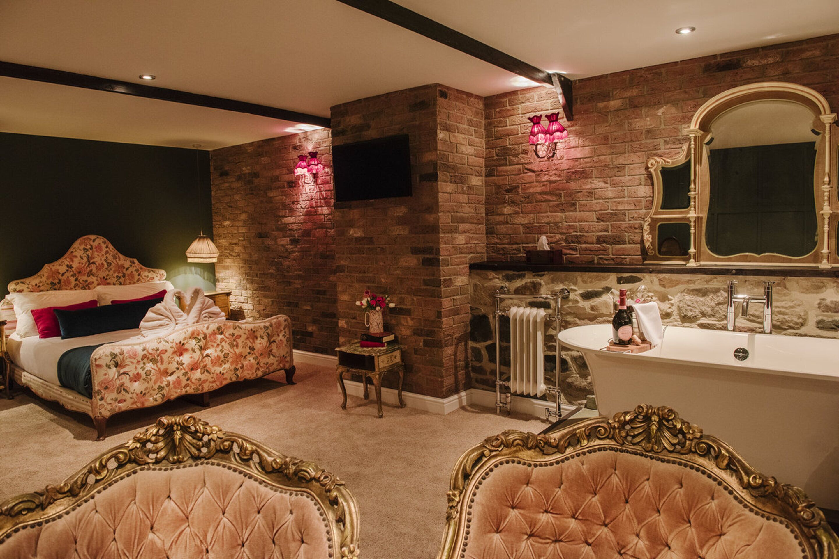 A bedroom with a brick wall, a bed with floral bedding, a vanity with a mirror, and a bathtub.