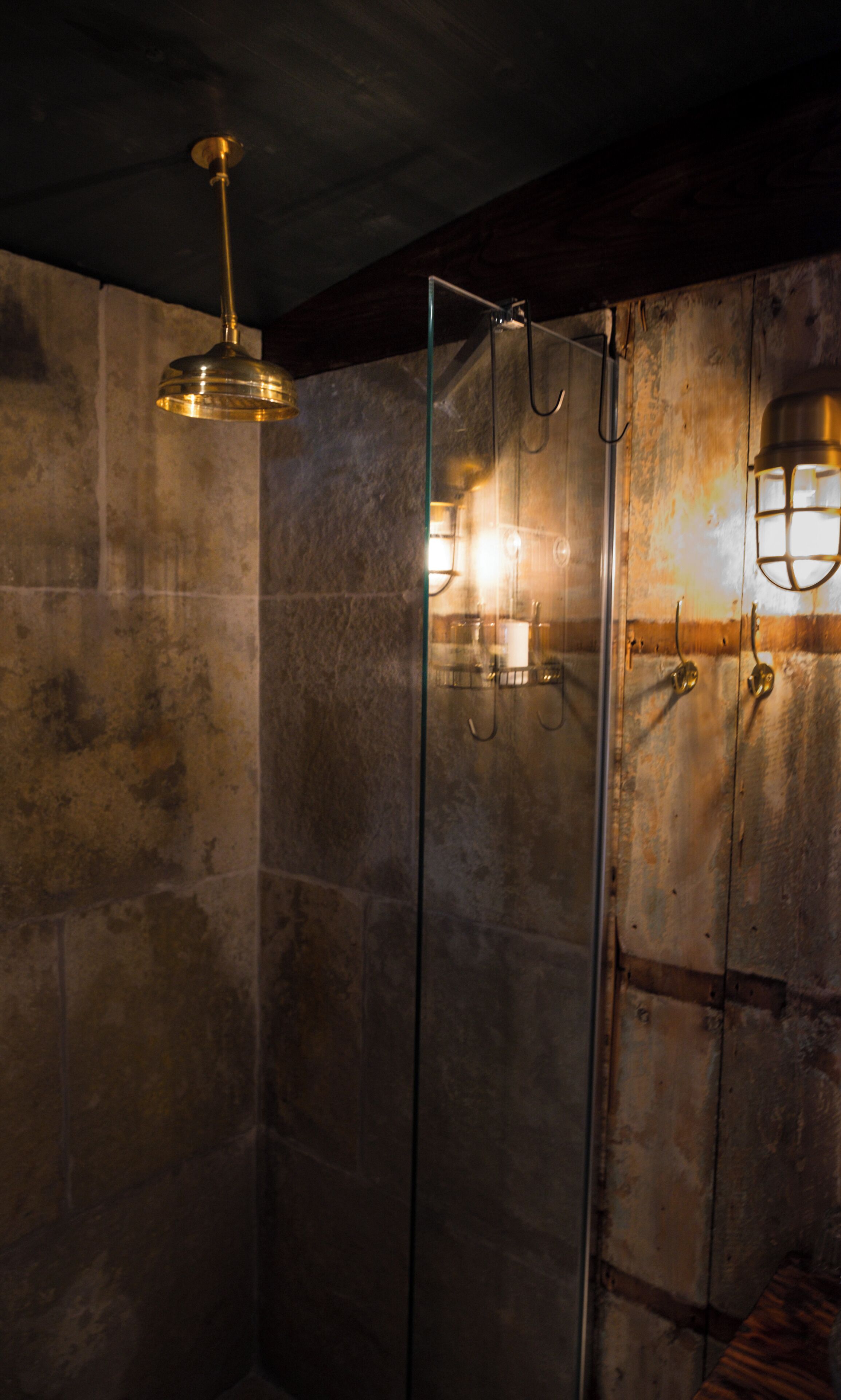 A glass-enclosed shower with a gold fixture, stone walls, and a wooden floor.