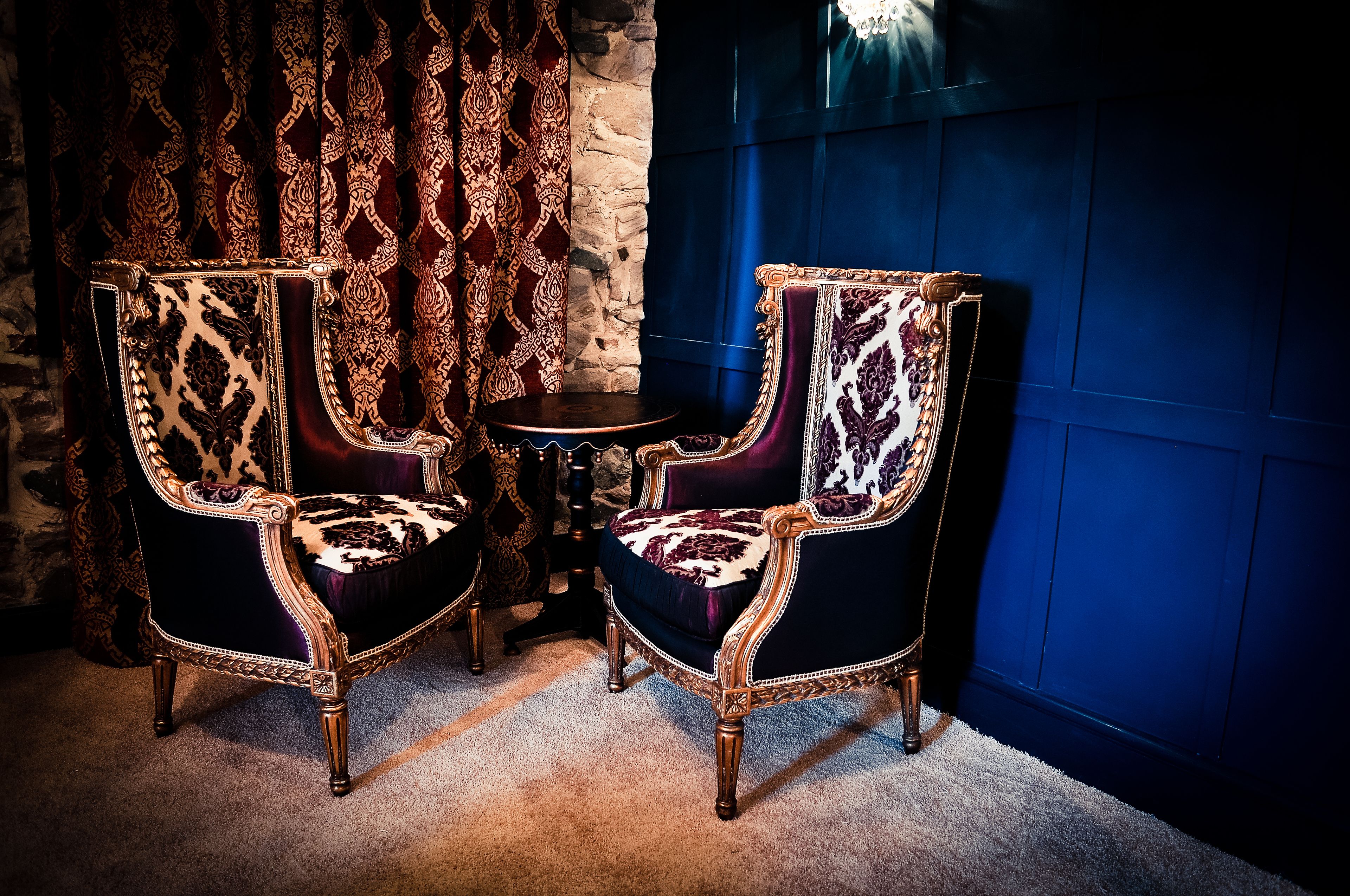 Two ornate chairs with intricate patterns, positioned next to a small round table in a room with a stone wall and blue paneled wall.