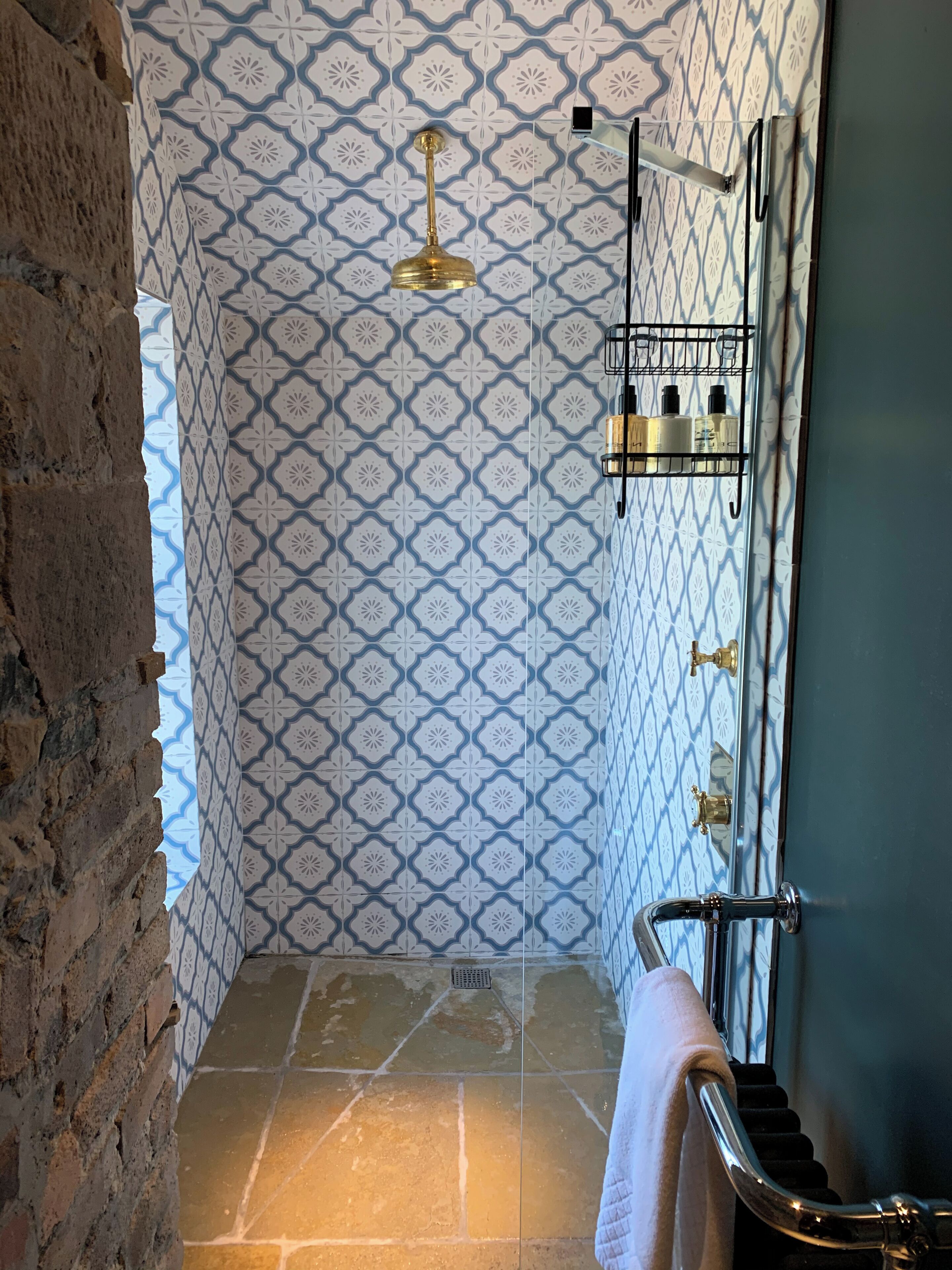 A bathroom with patterned tiles, a shower, a towel rack, and a brick wall.