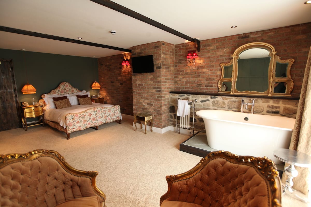 Une chambre avec un grand lit, un coin salon avec deux fauteuils, une grande baignoire et un mur en pierre.