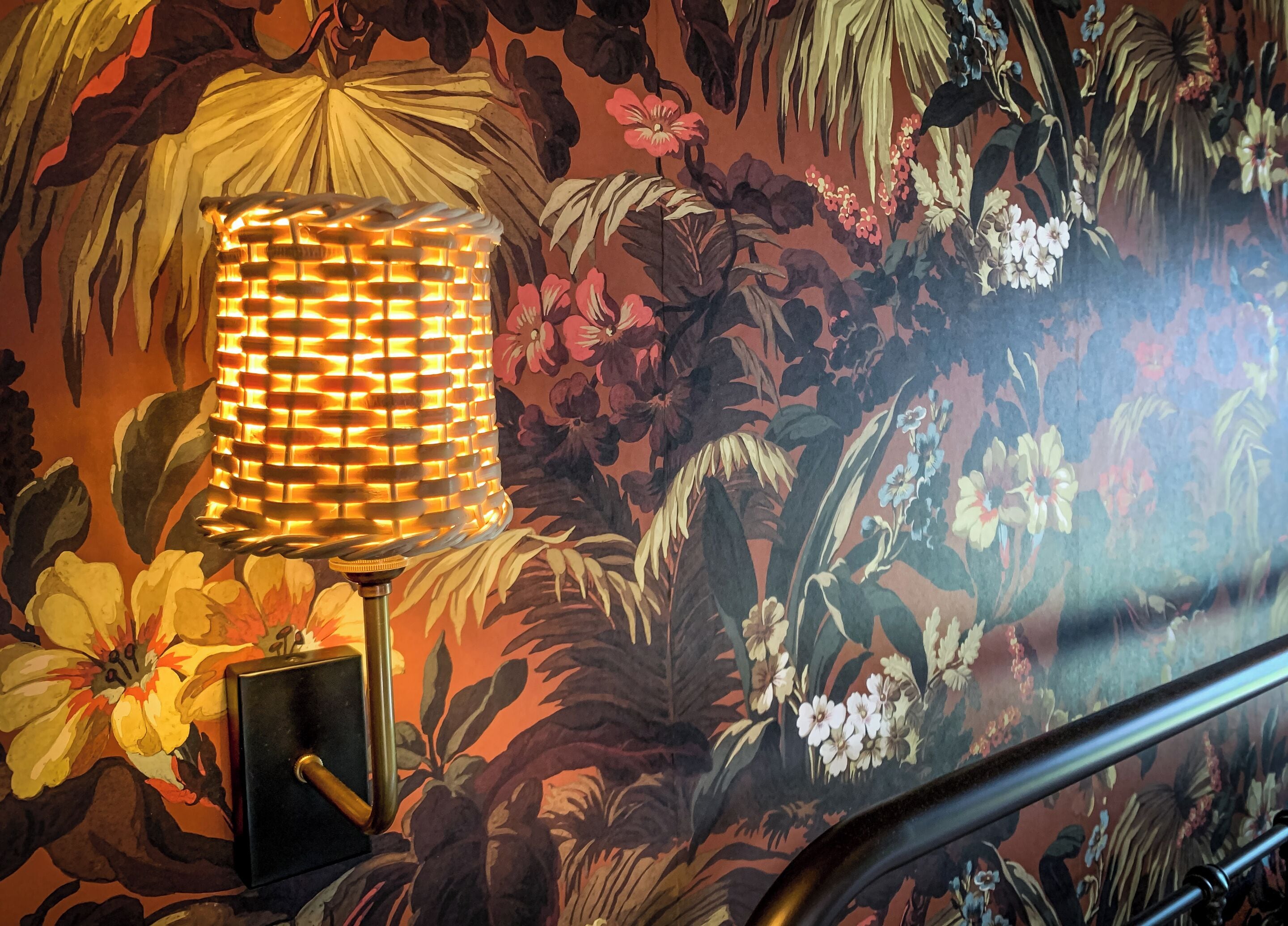 Una habitación con papel tapiz floral, una lámpara de pie y una barandilla de metal.