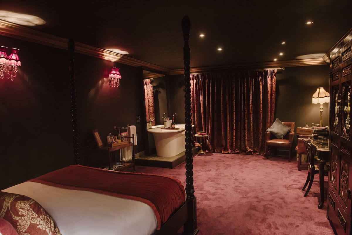 Un lit à baldaquin, une baignoire, un bureau et une chaise dans une pièce faiblement éclairée, avec des rideaux rouges.