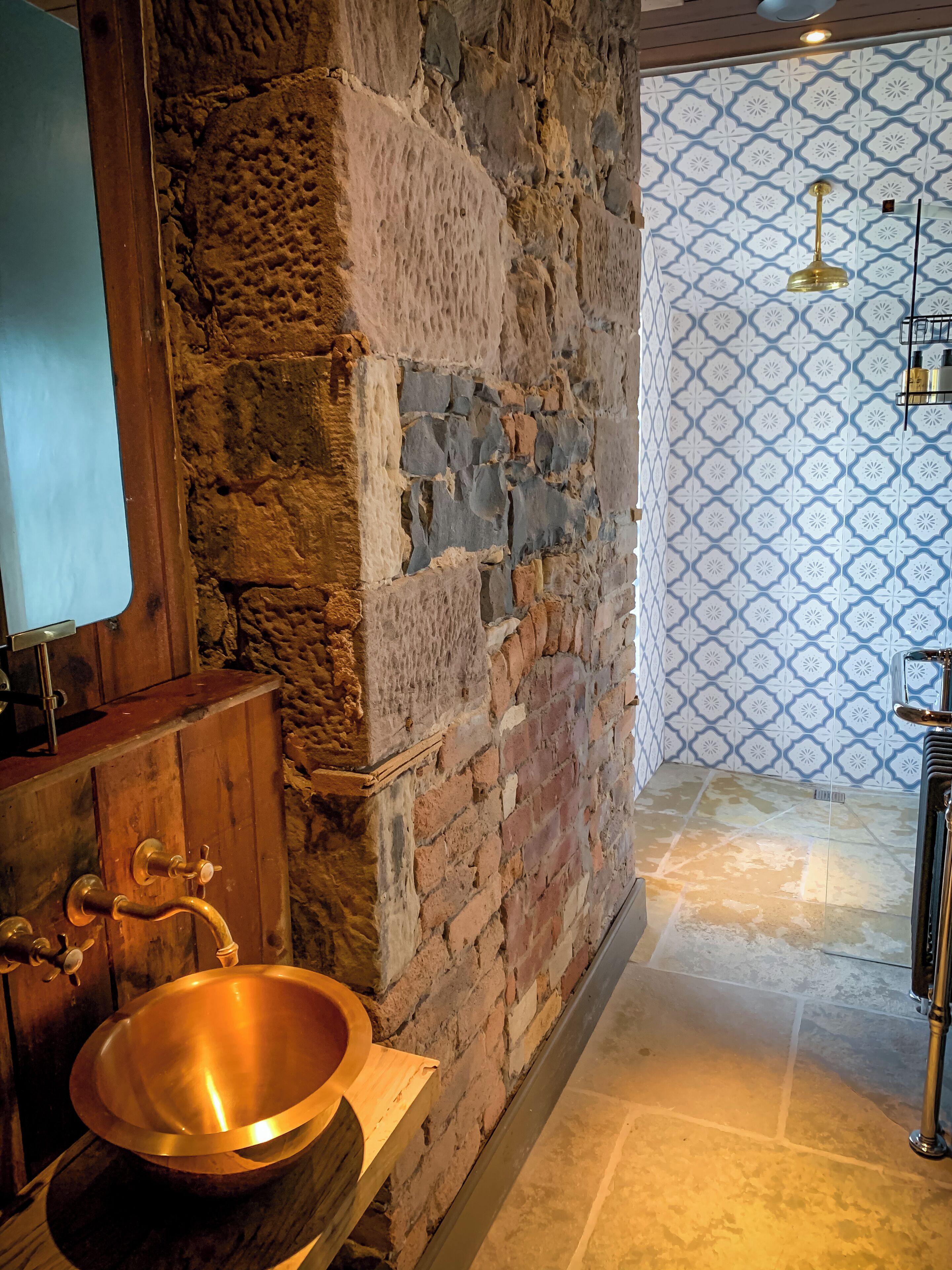 A bathroom with a stone wall, wooden vanity, copper basin, and patterned wall tiles.