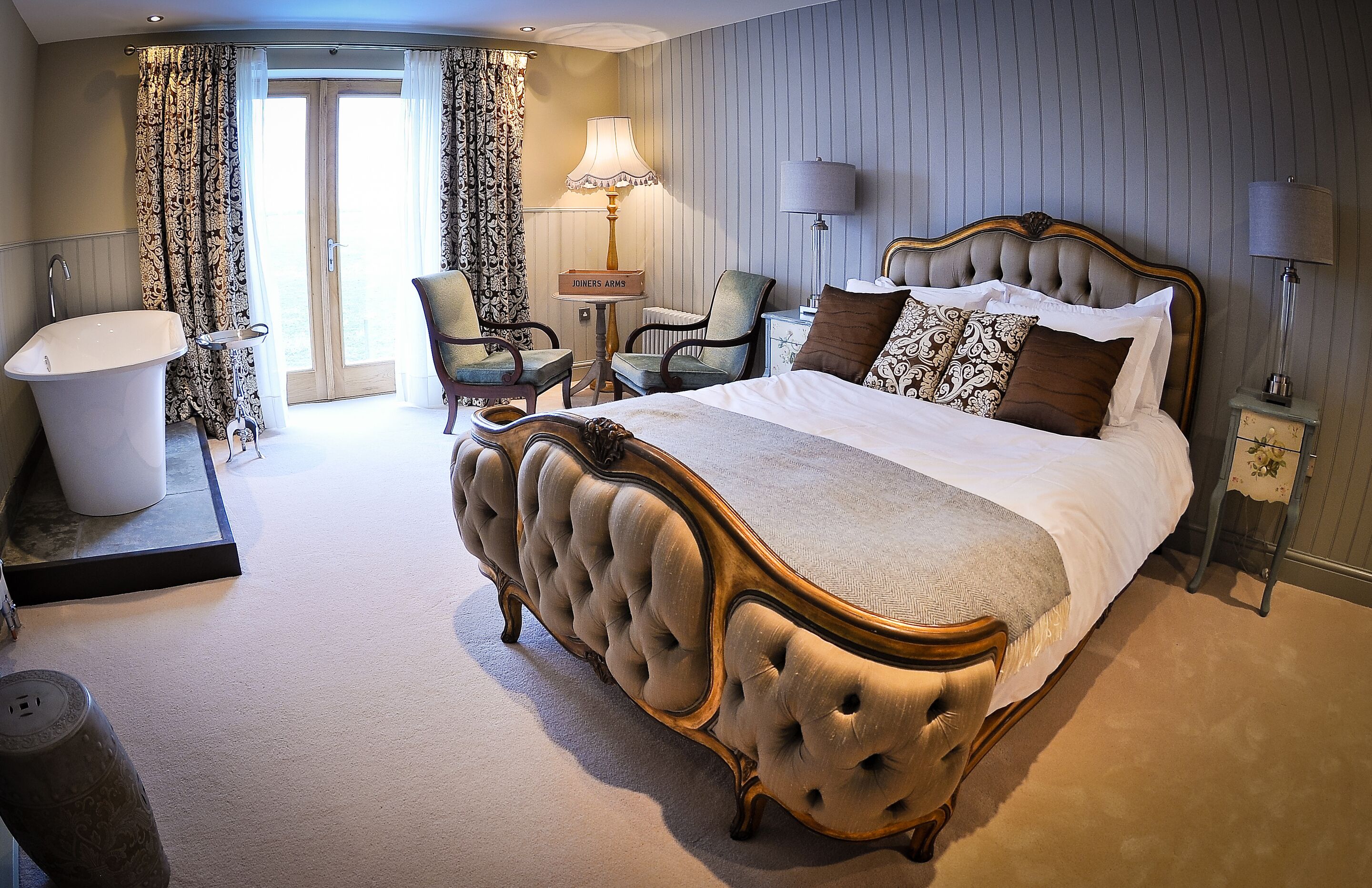 A spacious bedroom with a large bed, a sitting area with chairs, a vanity, and a bathtub.