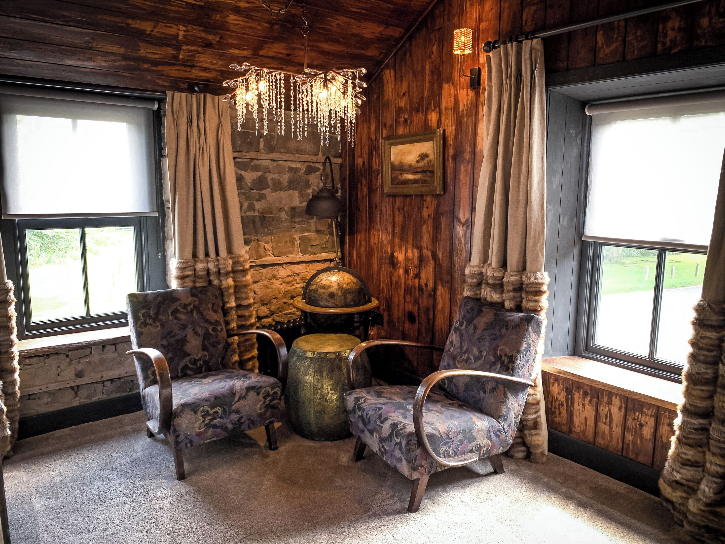 A rustic room with wooden walls, two patterned armchairs, a round table, a stone wall, and a hanging light fixture.