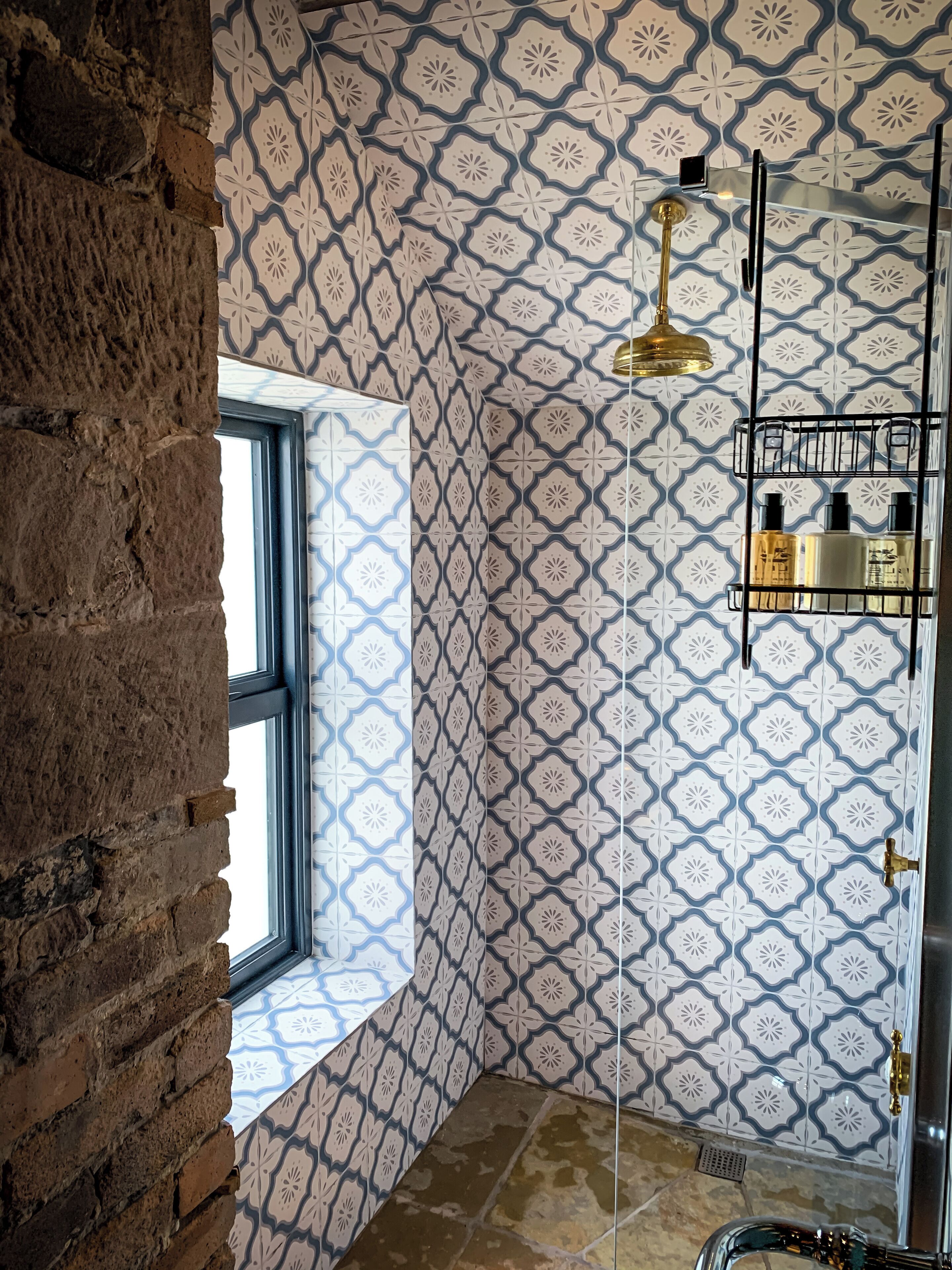 Une salle de bain avec des carreaux décorés, une fenêtre, une douche dotée d’une robinetterie dorée et une étagère contenant des articles de toilette.