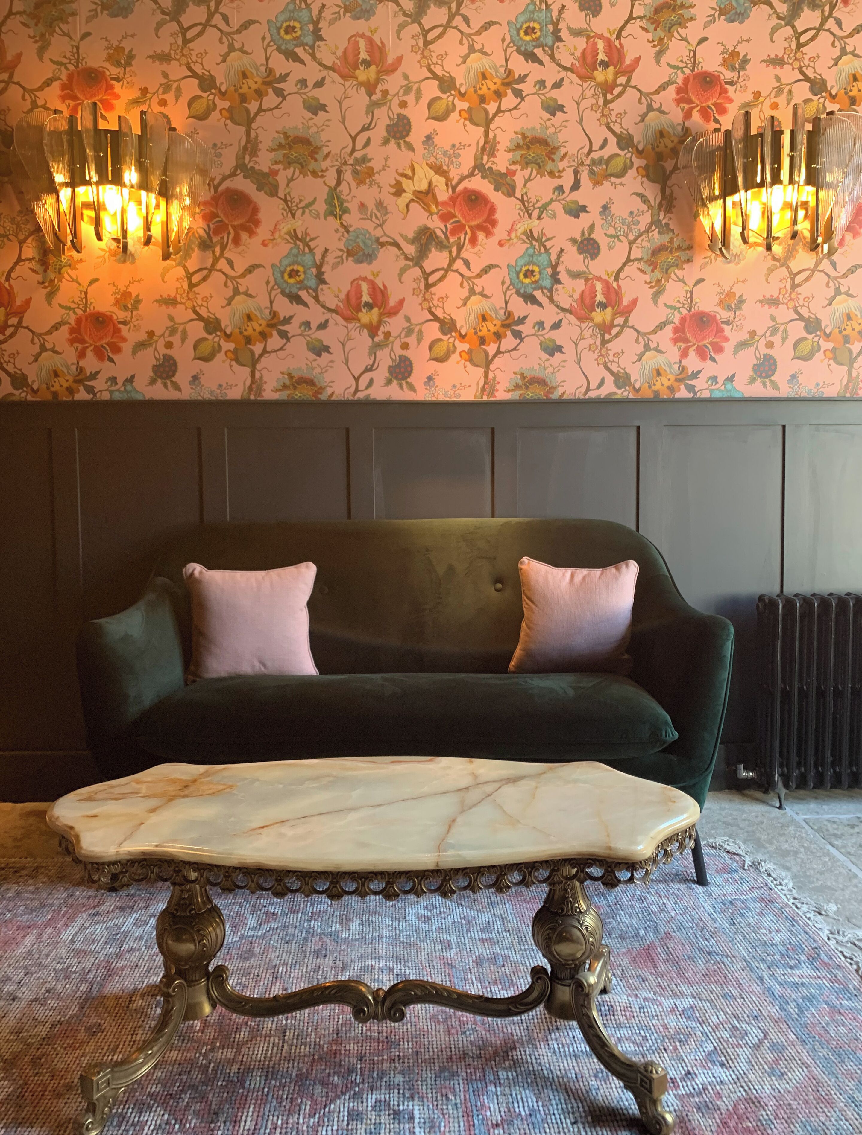 Un sofá verde oscuro con dos cojines rosa, una mesa de centro de mármol con patas ornamentales y un papel tapiz floral, con un radiador al fondo.