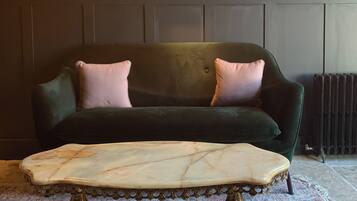 Un canapé vert foncé agrémenté de deux coussins roses, une table basse en marbre aux pieds décoratifs et un papier peint à motifs floraux, avec un radiateur placé derrière.
