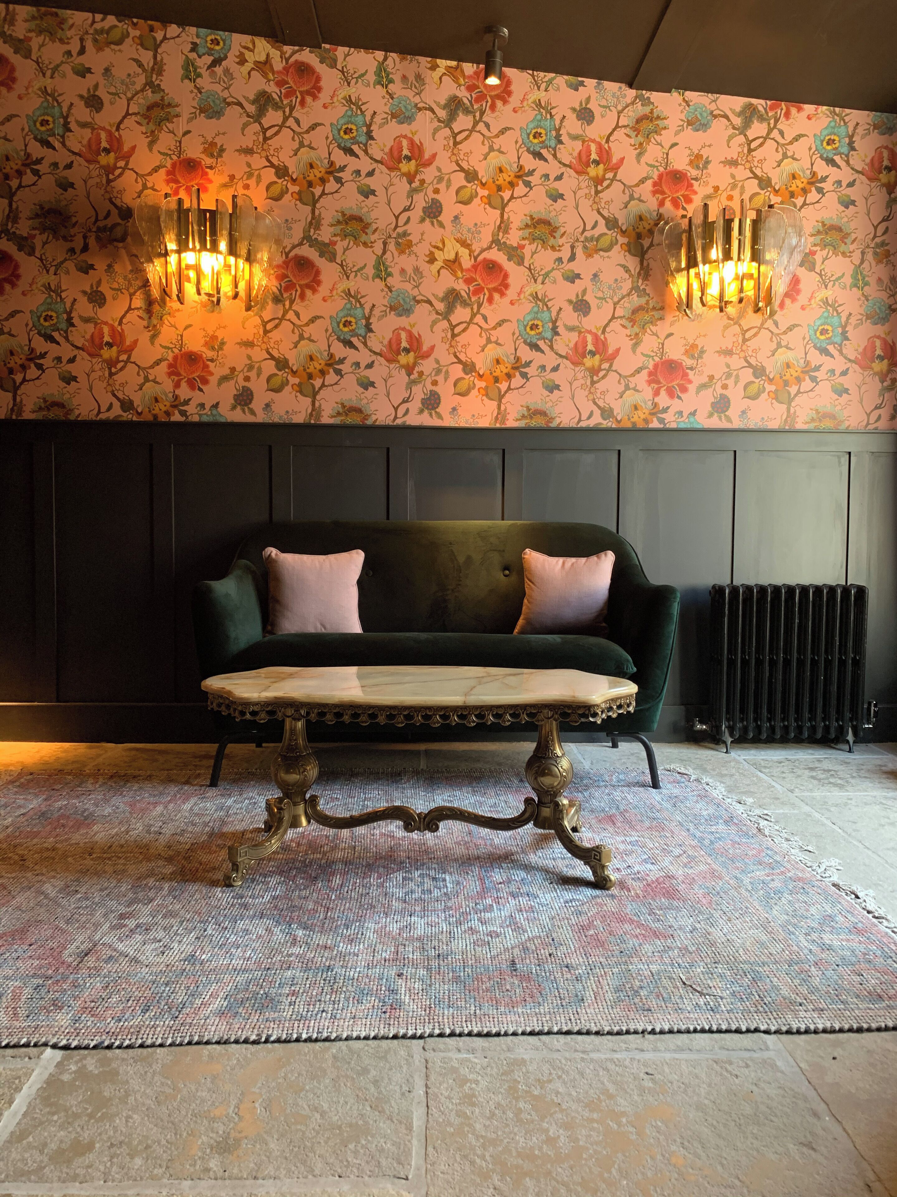 A room with a floral wallpaper, a dark green sofa with pink cushions, and a marble coffee table.