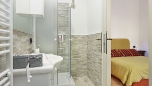 A bathroom with a glass shower enclosure, a white sink with toiletries, and a bed with a yellow bedspread. - B&B Pergola House (Lucca)