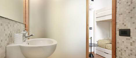A compact bathroom with a sink, toilet, and a small bedside table with a lamp.