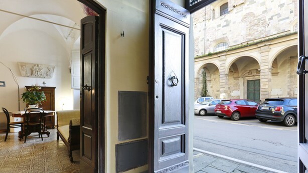 A hotel entrance with a dark wooden door, a tiled floor, and a view of parked cars and a building with arches. - B&B Pergola House (Lucca)