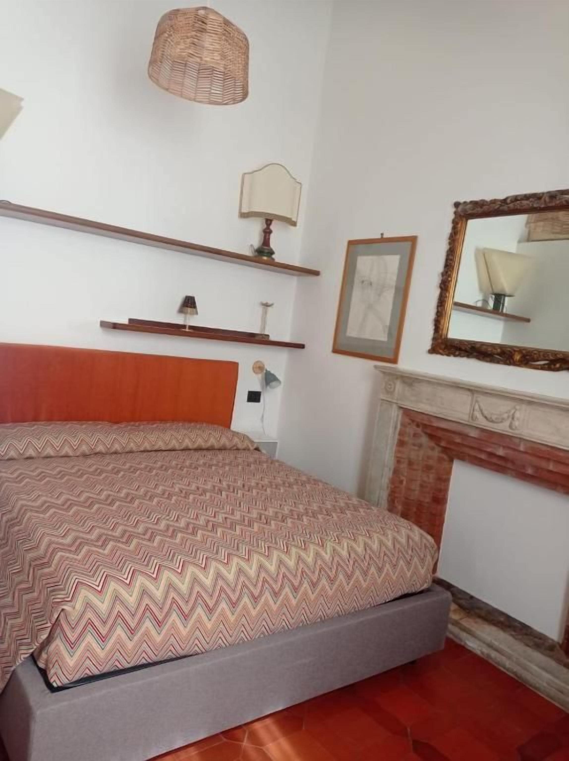 A bedroom with a bed, a fireplace, a mirror, and shelves with lamps.