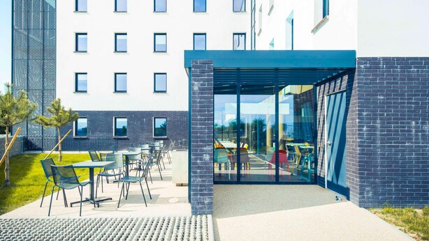 Ein modernes Gebäude mit überdachter Außenbestuhlung, einer Ziegel-Fassade und blauem Himmel.