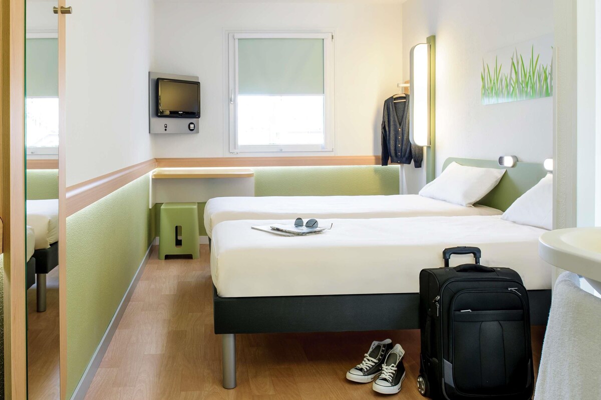 Une chambre d’hôtel avec deux lits, une télévision, une fenêtre et une valise.